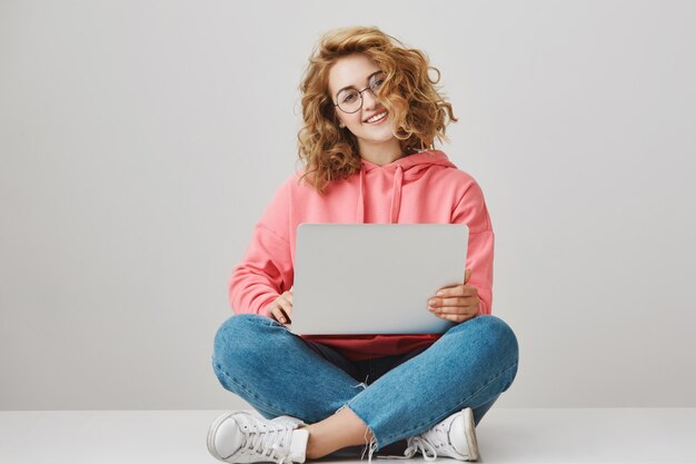 Cute freelance girl using laptop, sitting on floor and smiling