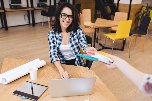 Carina giovane donna bruna amichevole in occhiali neri al tavolo prendendo libri e sorridendo al cliente. studiare all'università, lavorare come libero professionista, grande successo, grande squadra.