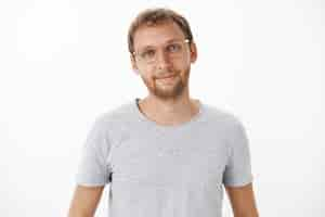 Free photo cute friendly and handsome male european with beard in glasses and gray t-shirt smiling happily with relaxed and relieved expression listening to customer question over white wall