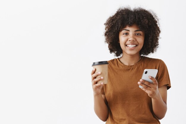 cute friendly-looking urban african american woman with afro hairstyle holding paper cup with coffee or tea and smartphone in hand smiling broadly reading news in morning