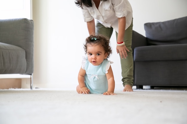 집에서 바닥에 크롤 링 귀여운 재미있는 아기. 작은 아이 뒤에 서있는 엄마. 부모와 어린 시절 개념