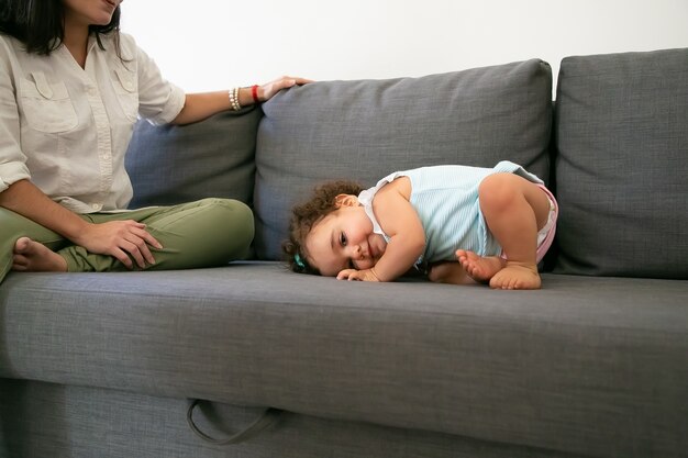 Cute funny baby girl in pale blue dress lying on grey couch near mom. Cropped shot. Parenthood and childhood concept