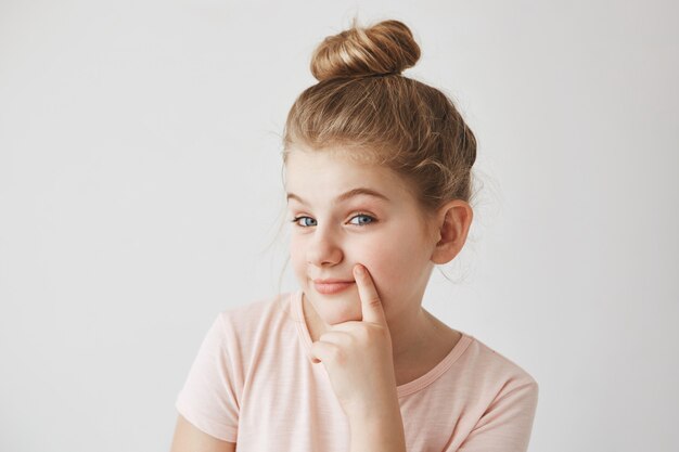 Cute funny blond little girl with bun hairdo holding finger near lips,  with raised eyebrows and sneaky face expression.