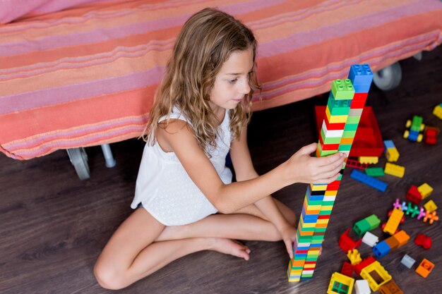 建設おもちゃのブロックで遊ぶかわいい面白いプレティーンの女の子の肖像画 カラフルなブロックに囲まれた木製の床に横たわって遊んでいる子供たち プレミアム写真