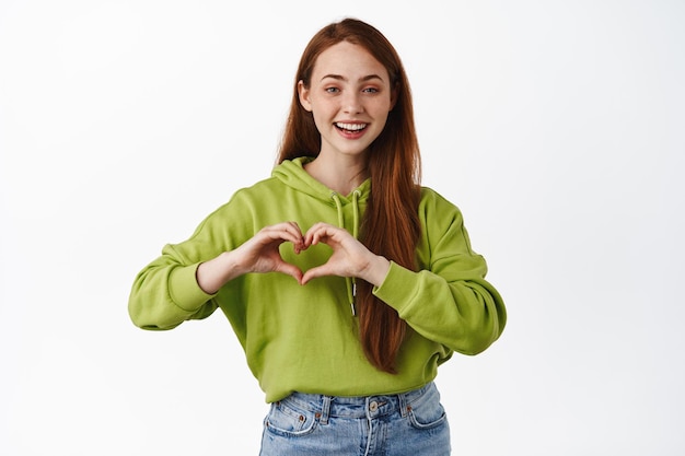 Cute ginger girl shows heart I love you sign, smiling and looking happy, adore something, feeling grateful, white background