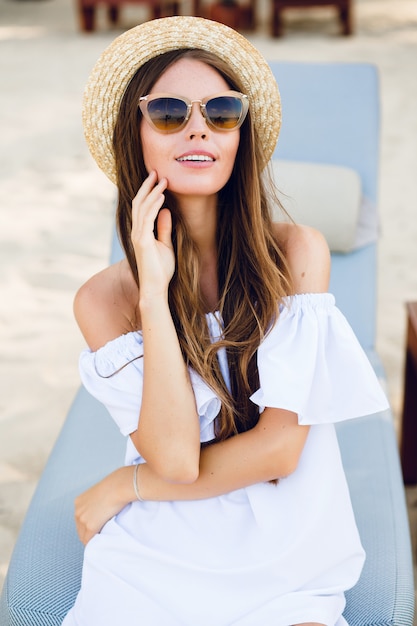 Cute girl in brown sunglasses and straw hat smiles widely and holds her hand near chin.
