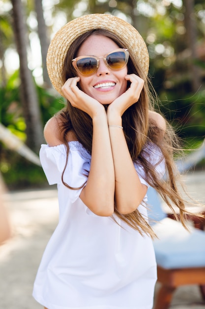 Cute girl in brown sunglasses and straw hat smiles widely and holds her hands near chin.