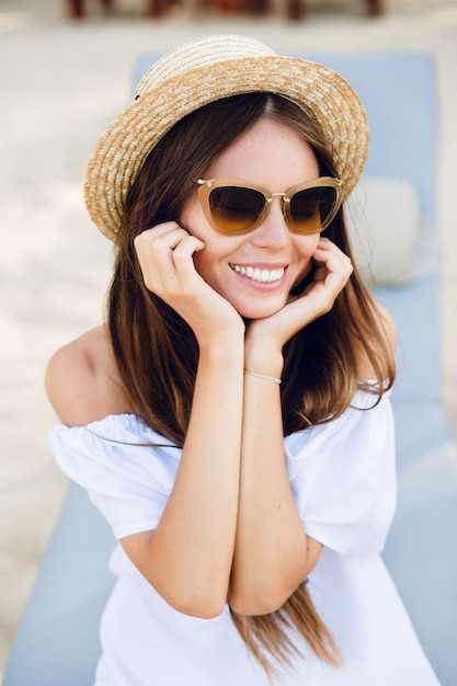 Ragazza carina in occhiali da sole marroni e cappello di paglia sorride ampiamente e tiene le mani vicino al mento.