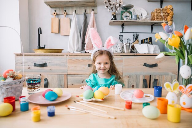 Cute girl in bunny ears sitting at table with Easter eggs