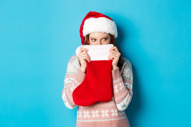 かわいい女の子は、クリスマスの靴下で顔を覆い、狡猾な視線で右を見つめ、サンタの帽子に立って、冬の休日を祝う、青い背景。