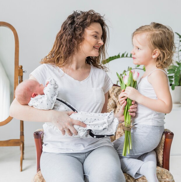 Cute girl giving tulip flowers to her mother holding baby sitting on arm chair