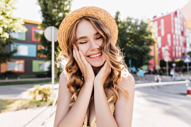 Cute girl in hat laughing with eyes closed on city. Romantic white woman dreamy posing in summer morning.