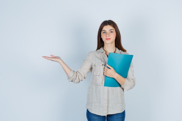 Cute girl holding folder, doing welcome gesture in shirt and looking gentle.