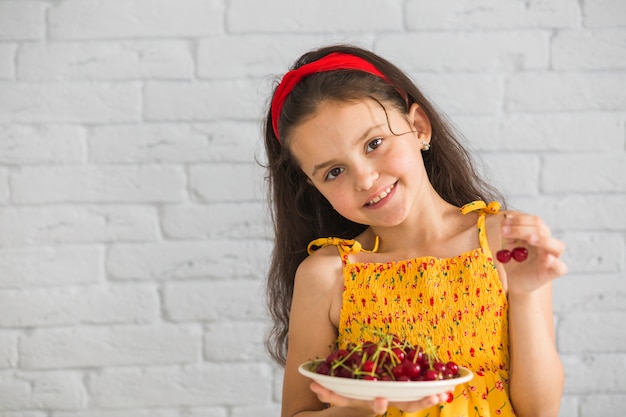 Free photo cute girl holding plate of red cherries against white brick wall