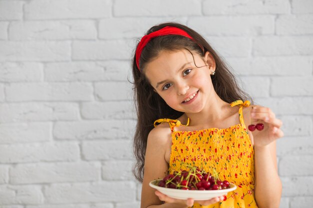 Cute girl holding plate of red cherries against white brick wall