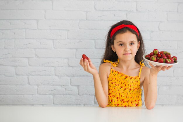 Cute girl holding plate of red strawberries against white brick wall