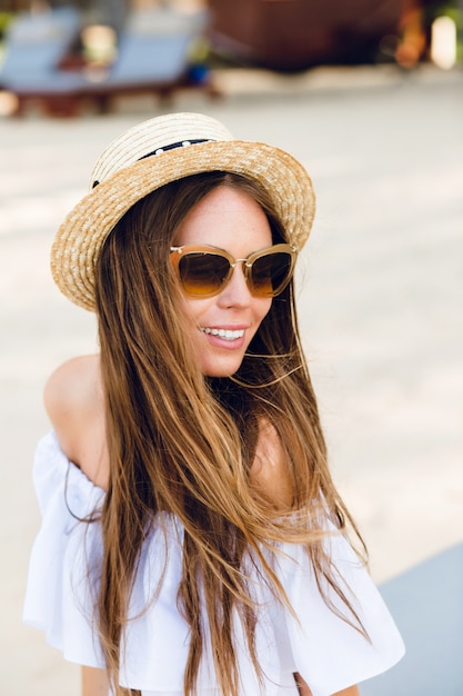 Милая девушка в коричневых очках и соломенной шляпе очаровательно улыбается.