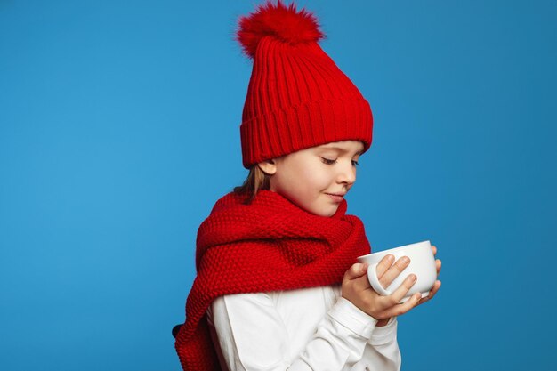 Милая девушка в вязаном красном шарфе и шляпе согревает руки, держа чашку