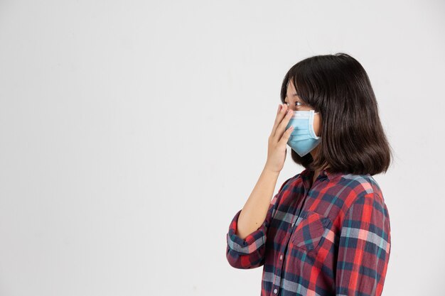 Cute girl is wearing mask and putting her hand up while close mouth by hand on white wall.