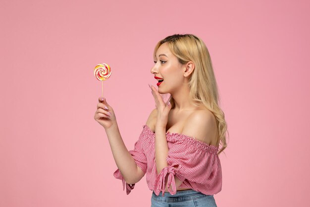 Cute girl lovely young lady with red lipstick in pink blouse looking at lollipop and smiling