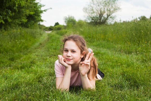 Cute girl lying on grassy land showing victory sign in beautiful nature