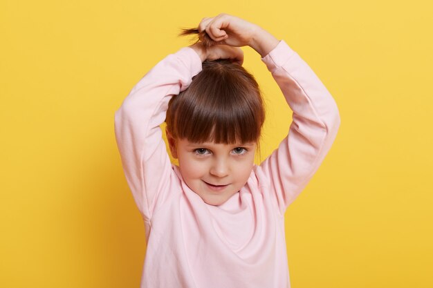 Cute girl making pony tail of her hair, raising her hands up, looks at camera, wearing pink casual sweater, standing isolated over yellow background, little charming female child makes hairstyle.