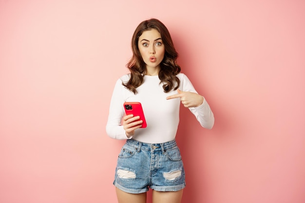 Симпатичная девушка, указывающая пальцем на смартфон с любопытным выражением лица, вы видели этот жест, показывающий предложение онлайн-покупок в приложении для мобильного телефона, розовый фон