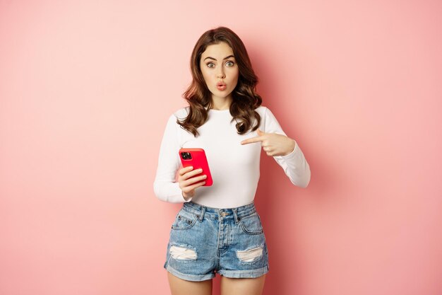 Cute girl pointing finger at smartphone with curious face expression, have you seen this gesture, showing online shopping offer on mobile phone app, pink background