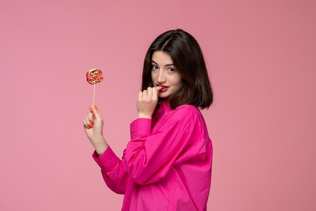 Симпатичная девушка, симпатичная молодая красивая брюнетка в розовой рубашке, кусающая палец леденцом на палочке