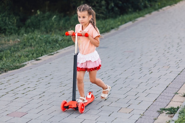 Cute girl riding red push scooter on walkway