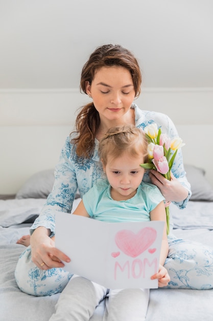 Cute girl sitting with her mother reading greeting card on bed