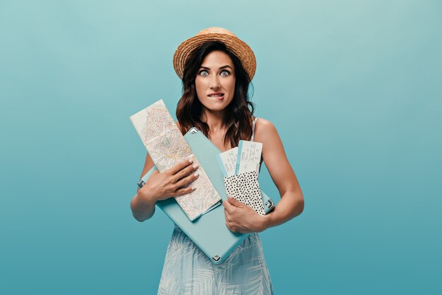 Cute girl in stylish sundress holds card, suitcase and tickets on blue background. Brunette in straw hat bites her lip and poses for camera.