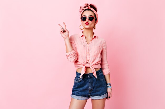 Cute girl in sunglasses in shape of heart blows kiss. Woman in headband, blouse and denim shorts showing peace sign.