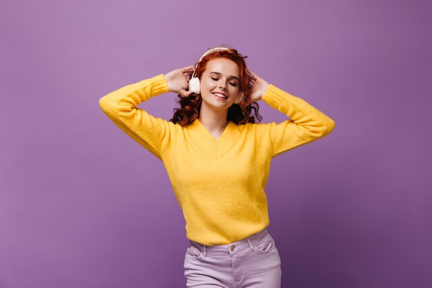 Cute girl in sweater listens to music in white headphones and dances on purple wall