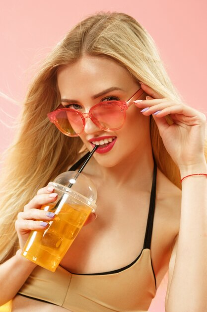 スタジオでポーズをとってオレンジジュースを飲む水着のかわいい女の子。ピンクの夏の肖像画白人ティーンエイジャー
