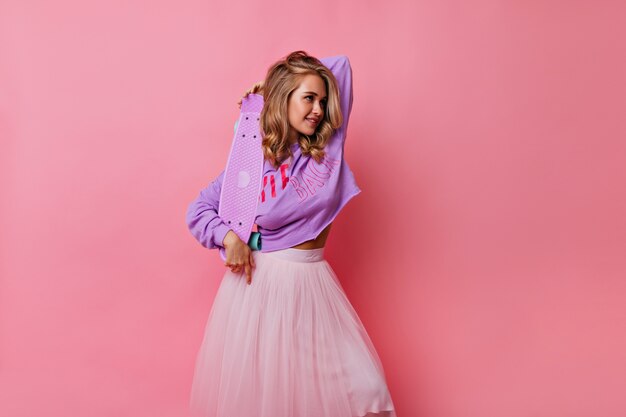 Милая девушка в модной повседневной одежде позирует со скейтбордом. Крытый портрет великолепной активной блондинки, изолированной на розовом.