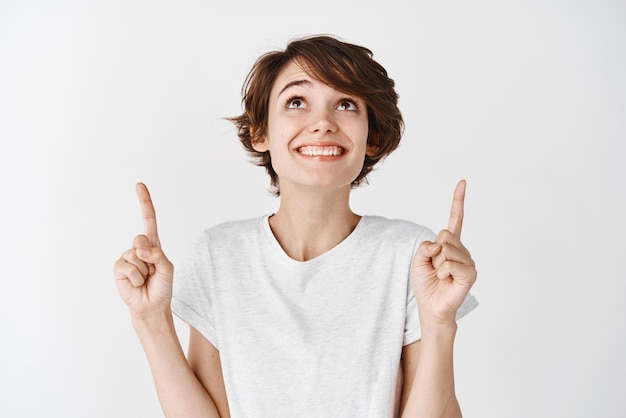 Cute girl in tshirt pointing looking up with dreamy smile staring at promo offer standing against white background