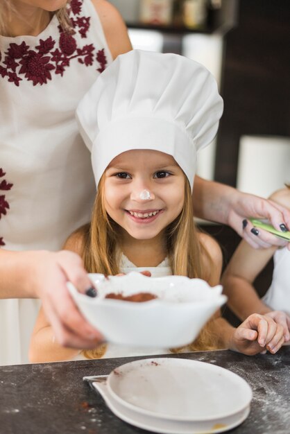 台所で料理をしながら前の母に立っているシェフの帽子をかぶっているかわいい女の子