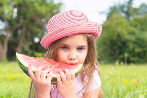 Cute girl wearing pink hat eating watermelon slice in the park