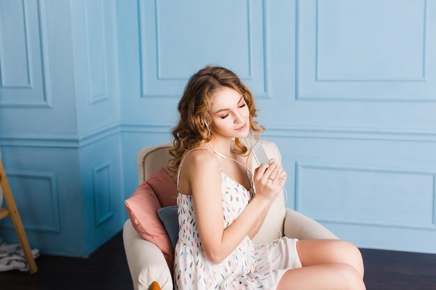 Cute girl with blond curly hair sits on armchair in studio with blue walls with eyes closed. She wears white dress and shoes. She listens to music on smartphone and earphones and looks dreamy.