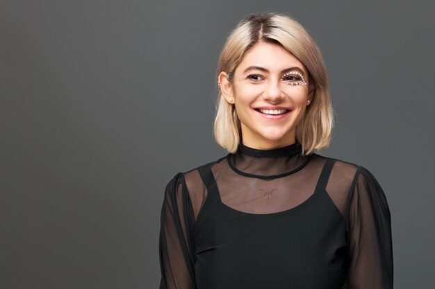 Cute girl with dyed bob hairstyle, artistic make up and nose ring wearing transparent dress over black strap top, posing against blank wall  with copy space for your information