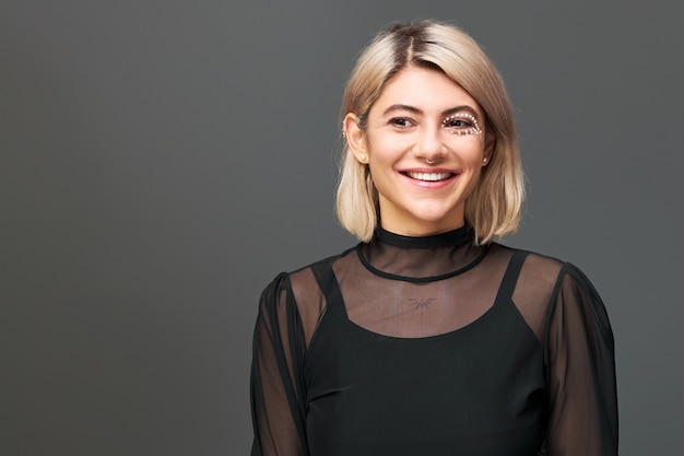 Cute girl with dyed bob hairstyle, artistic make up and nose ring wearing transparent dress over black strap top, posing against blank wall  with copy space for your information