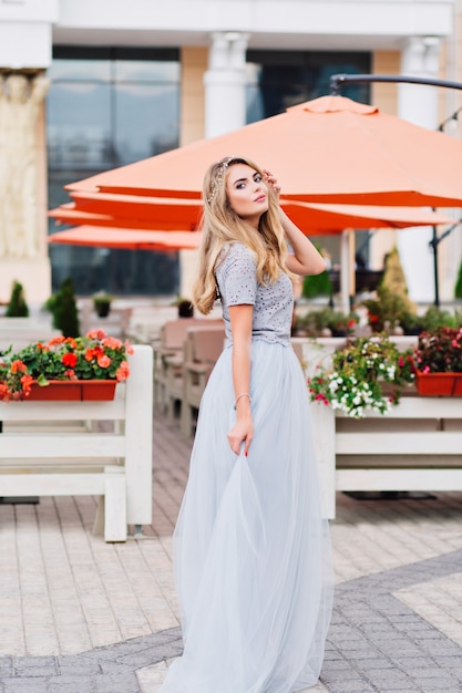 Cute girl with long blonde hair in long blue tulle skirt is walking on street.