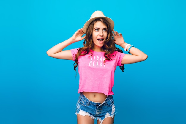 Free photo cute girl with long curly hair in hat posing on blue background in studio. she wears shorts, pink t-shirt. she looks wondered.