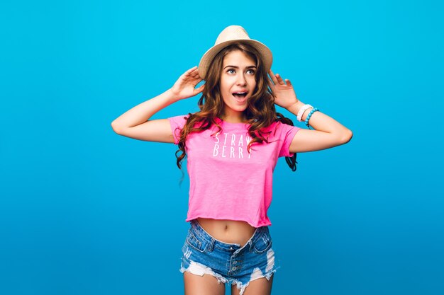スタジオで青い背景にポーズの帽子で長い巻き毛を持つかわいい女の子。彼女はショートパンツ、ピンクのTシャツを着ています。彼女は不思議に見えます。