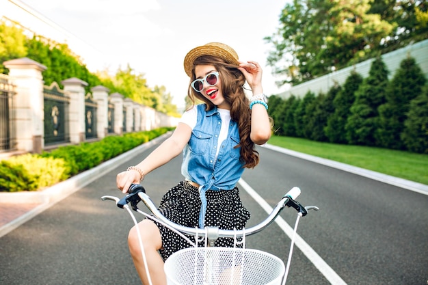 無料写真 サングラスをかけた長い巻き毛のかわいい女の子が自転車で道路のカメラに向かっています。彼女はロングスカート、ジャーキン、帽子をかぶっています。彼女は幸せそうだ。