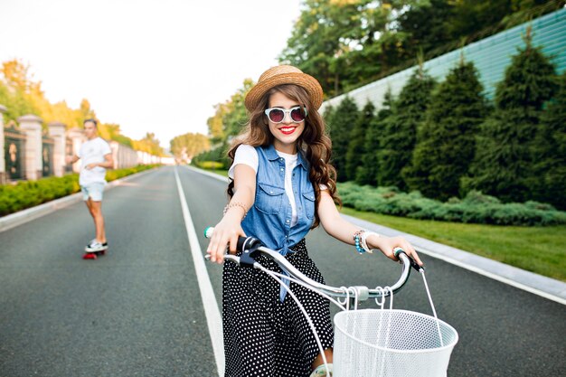 道路上のカメラに自転車を運転するサングラスで長い巻き毛を持つかわいい女の子。彼女はロングスカート、ジャーキン、帽子をかぶっている。ハンサムな男は背景にスケートボードに乗っています。