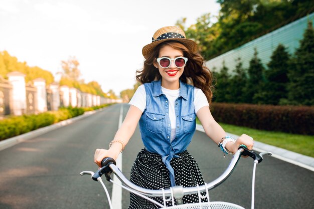 道路上のカメラに自転車を運転するサングラスで長い巻き毛を持つかわいい女の子。彼女はロングスカート、ジャーキン、帽子をかぶっている。彼女は幸せそうです。