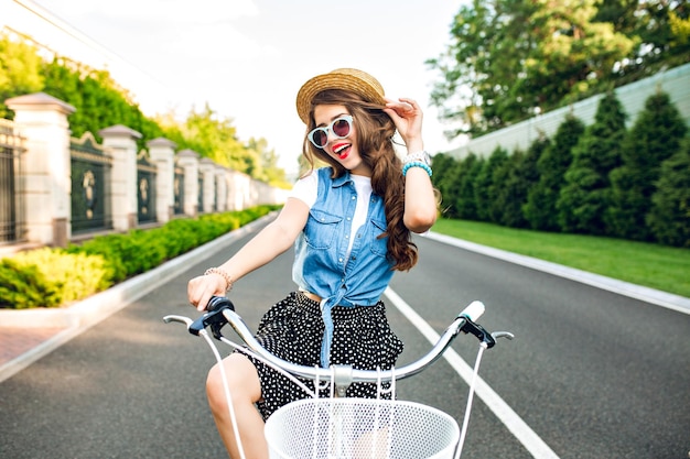 Cute girl with long curly hair in sunglasses is going by bicycle to camera on road. she wears long skirt, jerkin, hat. she looks happy.