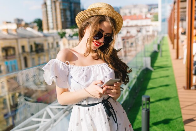 Cute girl with long hair in sunglasses is standing on the terrace. She wears a white dress with bare shoulders, red lipstick and hat . She is typing on phone.